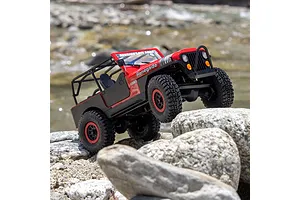 Axial SCX30 Jeep CJ-7 Renegade RC rock crawler in red navigating rocky terrain outdoors near water.