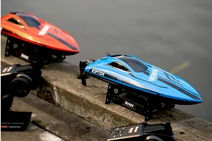 Blue Atrek Riptide RC speed boat and transmitter on a wooden surface with a red boat in the background.