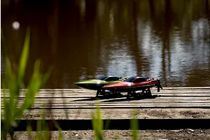 Two Atrek Rogue RC speed boats, one orange and one green, sitting on display stands on a wooden dock by the water.