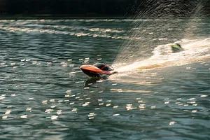 An orange Atrek Rogue RC speed boat speeding across a lake, kicking up a large spray of water in the sunlight.