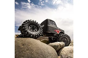 Matte black RC crawler with cab-only body and shark mouth decal driving over large rocks outdoors under a cloudy sky.