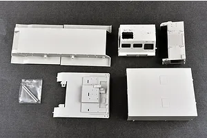 Unassembled grey plastic model kit parts for a military vehicle, including chassis and cabin components, laid out on a dark grey surface.