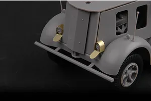 Close-up of the front end of an unpainted grey plastic model half-track vehicle with brass headlight covers.