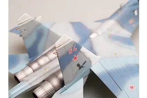 A close-up, high-angle view of a scale model Su-27 fighter jet, showing the tail fins with red number 64 and a red star emblem.