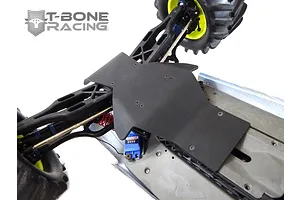 A black T-Bone Racing skid plate installed on the chassis of a Traxxas E-Maxx RC truck, showing the front suspension and steering servo.