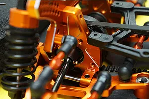 Close-up of an orange anodized aluminium RC car suspension assembly with black plastic components and a drive belt.