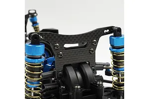 A close-up view of a carbon fibre front shock tower installed on an RC car chassis, featuring blue anodized shock absorbers and gold springs.