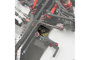 A close-up view of a 10g Yeah Racing black brass chassis balancing weight installed on a carbon fiber RC car chassis.
