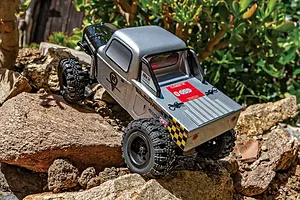 Element RC Enduro24 Ecto silver RC rock crawler driving over rough outdoor rocks in a garden setting.