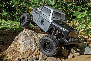 Element RC Enduro24 Ecto 1/24 scale RC rock crawler in silver and black, climbing over a rock in an outdoor garden setting.