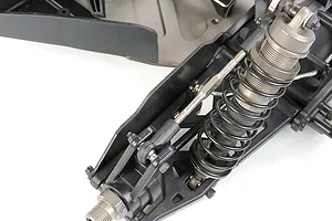 A close-up view of a carbon fibre rear suspension arm guard installed on an RC car chassis, featuring a coil-over shock absorber and suspension linkage.