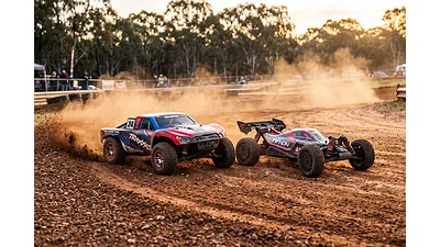 A Traxxas stadium truck and an ARRMA buggy racing side-by-side on a dusty dirt track under golden sunlight.