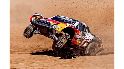 A Traxxas Slash short course RC truck performing a wheelie on a dirt track, kicking up dust.