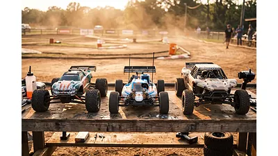 Three RC cars representing electric, nitro, and petrol power types lined up on a pit bench at an outdoor dirt track.