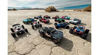 A large collection of various Arrma RC trucks and buggies arranged on a dry, cracked desert ground under a blue sky.