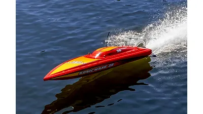 A red and yellow remote control speed boat racing across the water, kicking up a large spray of water from the stern.