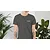 A person wearing a dark grey T-shirt with a small TDK logo on the chest, standing between two indoor plants.