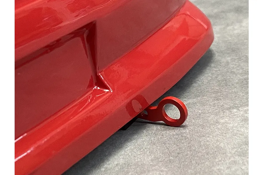 A close-up view of a red metal tow hook attached to the front bumper of a glossy red RC car body shell.