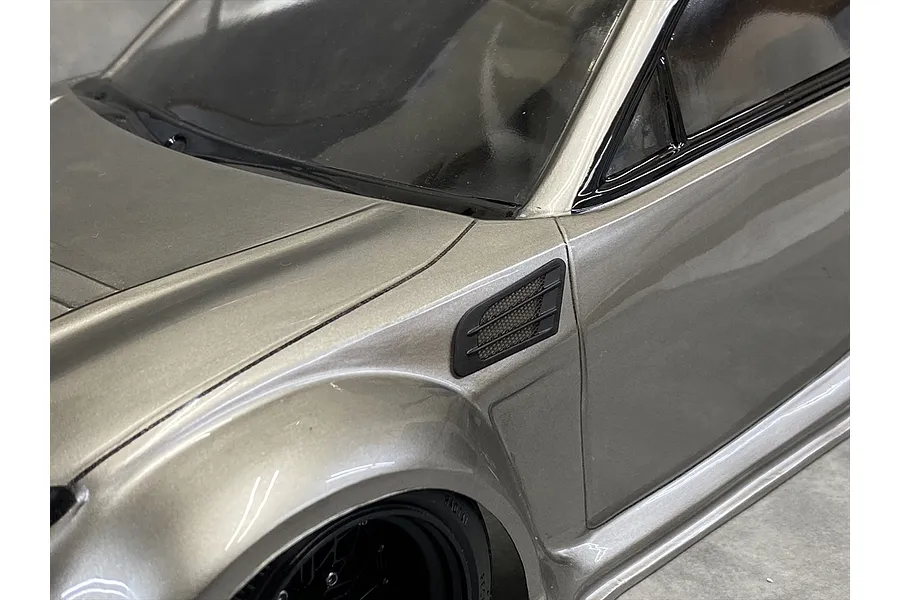Close-up of a silver RC car body showing a black fender duct accessory installed on the front quarter panel.