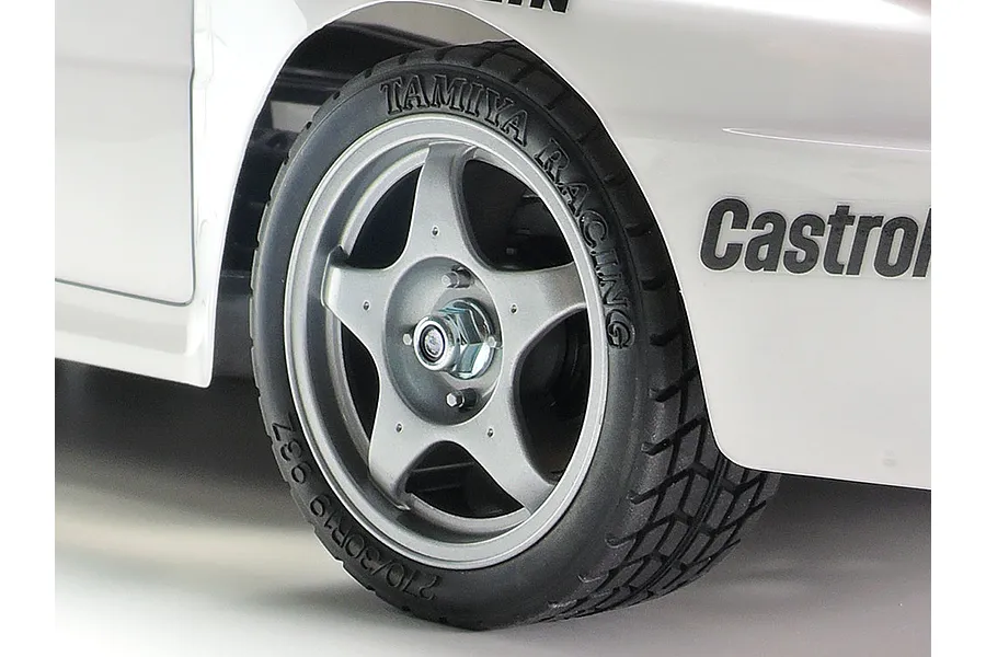 Close-up of a Tamiya RC car wheel with a five-spoke silver rim and treaded rubber tire on a white body shell with Castrol branding.