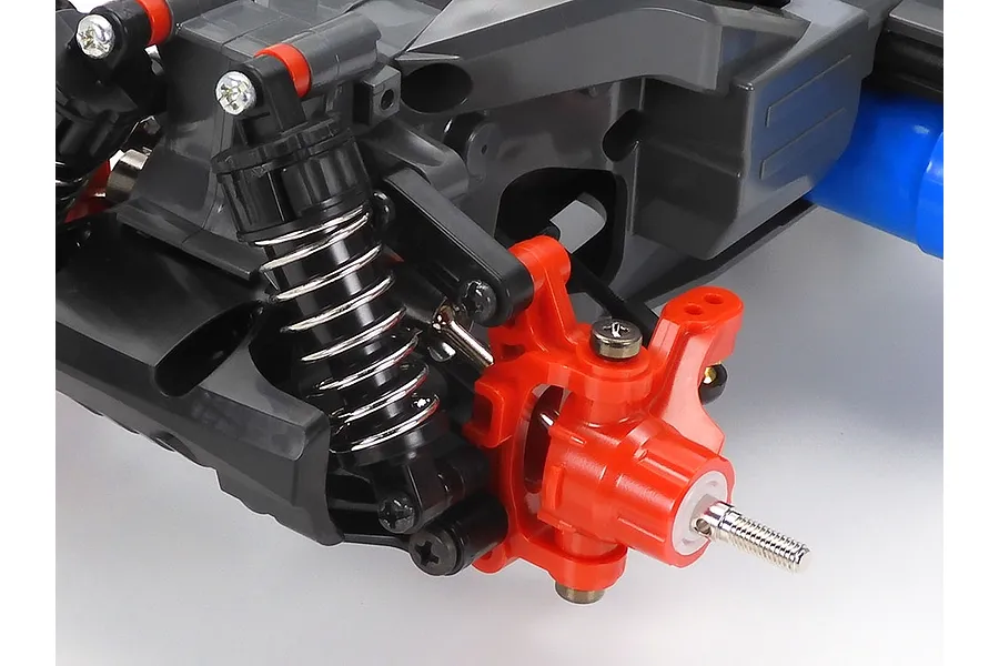Close-up of the front suspension and steering knuckle assembly on a Tamiya MF-01X RC car chassis, featuring a coil-over shock and bright orange plastic parts.