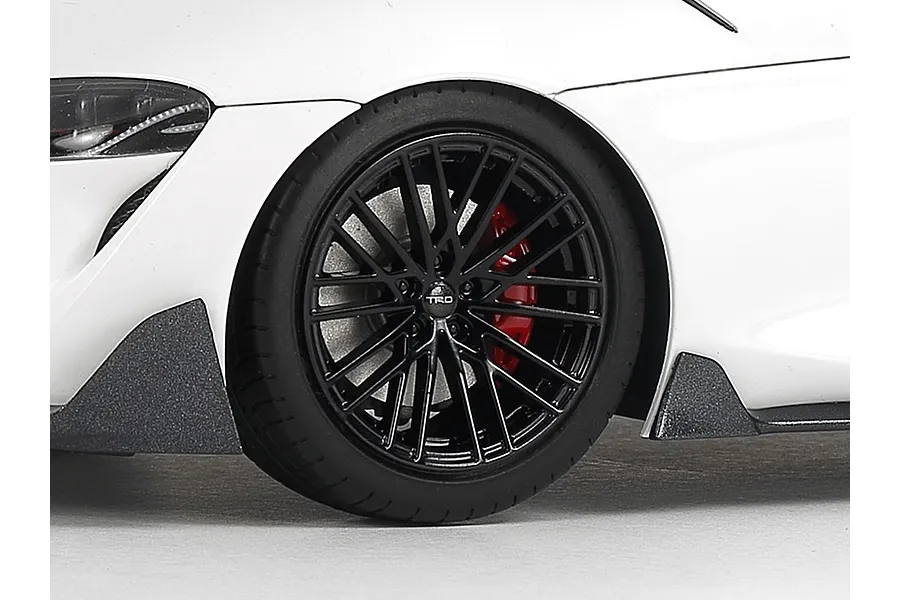 Close-up of the front wheel and fender of a white Tamiya 1/24 Toyota GR Supra scale model car featuring black TRD rims and red brake calipers.