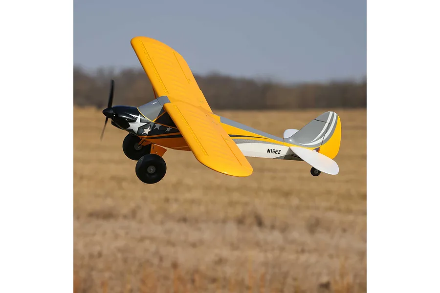 A HobbyZone Carbon Cub S 2 RC plane flying over a field, featuring a yellow, grey, and black paint scheme with star decals.