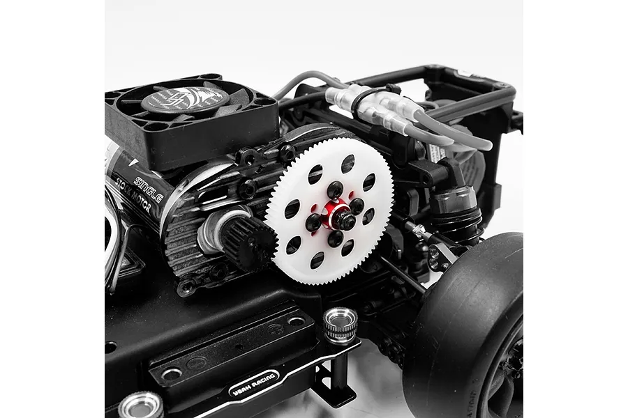 Close-up of a red aluminium spur gear mount installed on an RC car chassis with a white spur gear and black motor cooling fan.