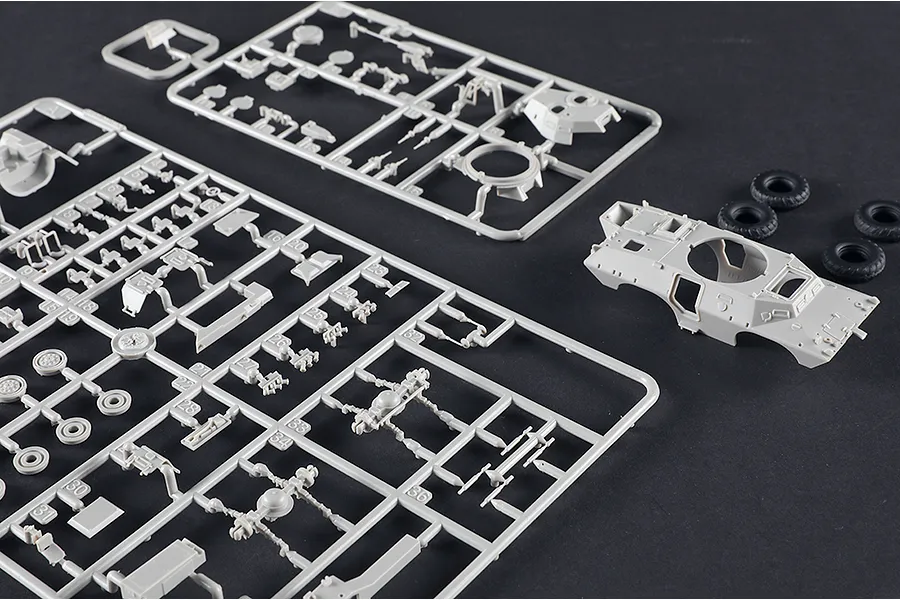 Plastic model kit sprues and a vehicle hull for a V-150 Commando APC, showing grey plastic parts and four black rubber tires on a dark surface.