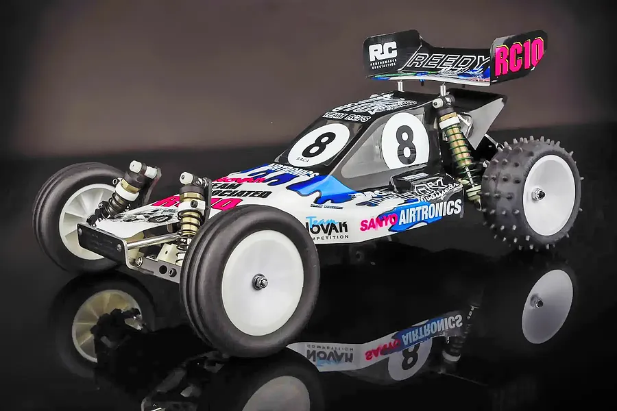 A classic Team Associated RC10 Kinwald Edition 1/10 scale RC buggy with white wheels, black tires, and a detailed racing livery on a reflective surface.