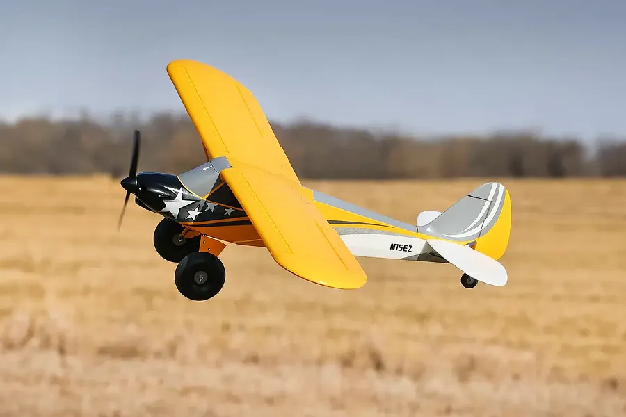 A yellow and grey HobbyZone Carbon Cub S 2 RC plane flying over a field.