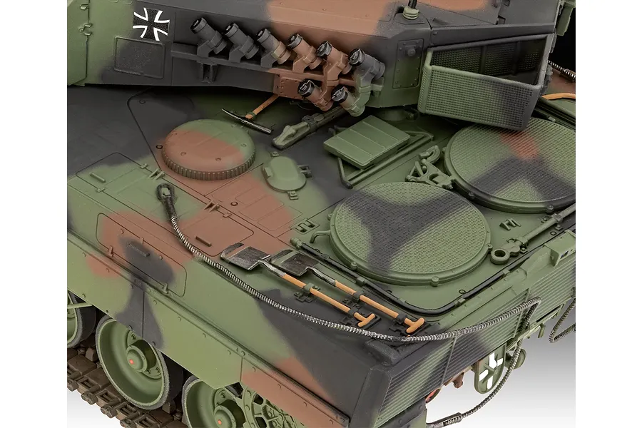 Close-up of a 1/35 scale Leopard 2A6M+ model tank showing detailed camouflage paint, turret smoke launchers, and tools mounted on the hull deck.