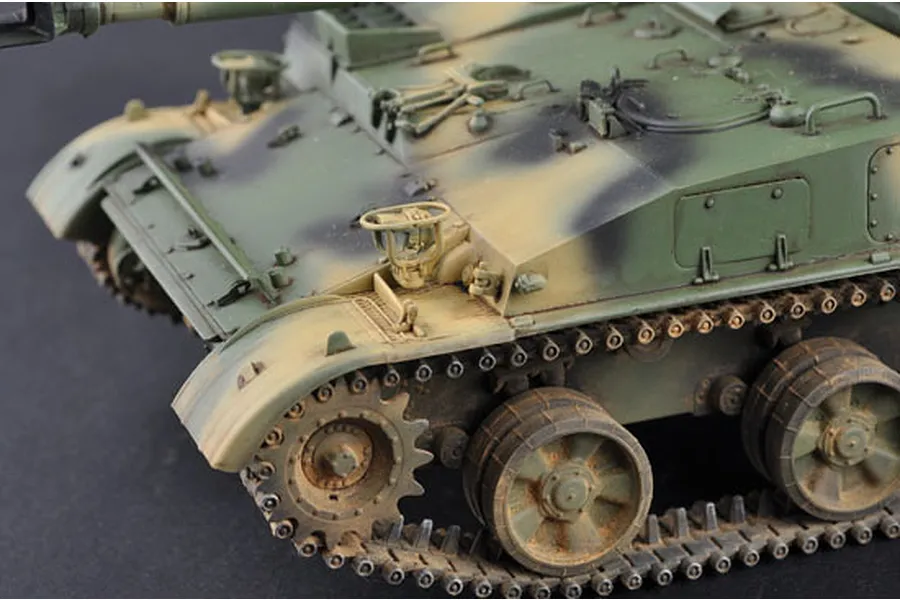 A close-up view of a 1/35 scale model of a Soviet 2S3 self-propelled gun, showing detailed weathering, camouflage paint, and tracks.