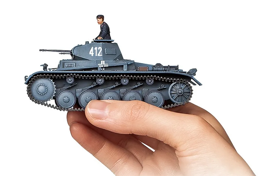 A hand holding a small, assembled 1/48 scale Tamiya German Panzer II tank model painted in grey with a tank commander figure.