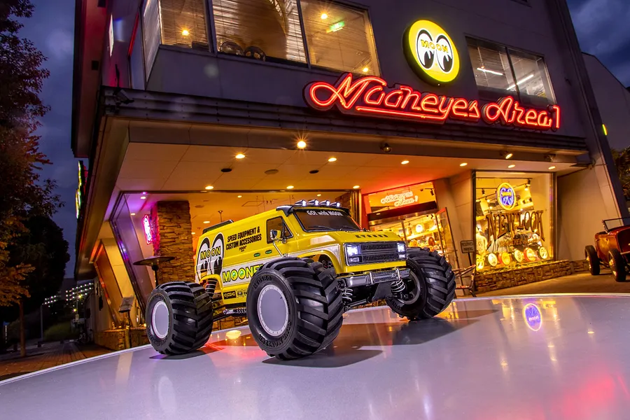 A yellow Kyosho Mad Van RC monster truck with Mooneyes branding parked on a reflective surface in front of a Mooneyes store at dusk.