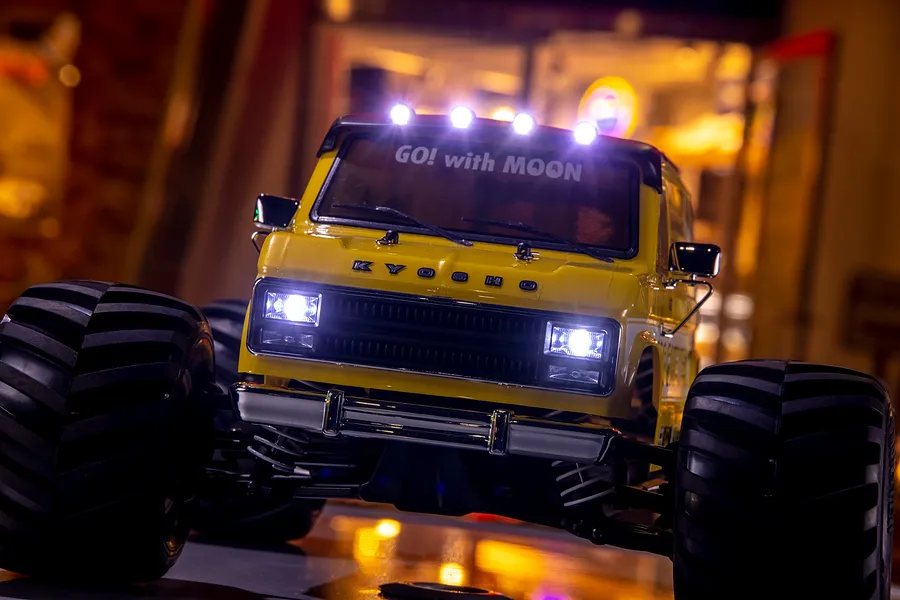 Front view of a yellow Kyosho Mad Van RC monster truck with Mooneyes branding, featuring illuminated headlights and roof lights in a dimly lit setting.