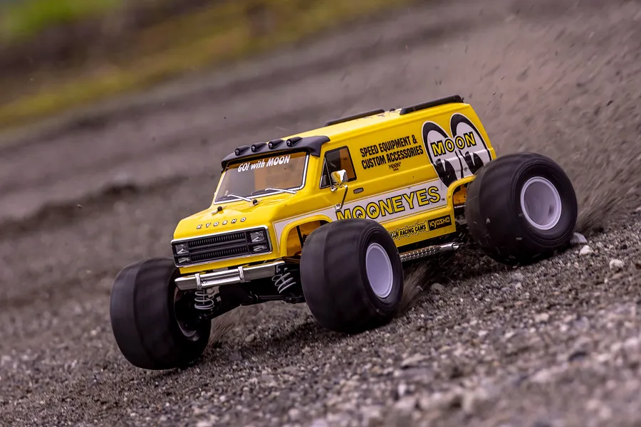 A yellow Kyosho Mad Van RC monster truck with Mooneyes branding driving fast on a gravel surface, kicking up dirt.