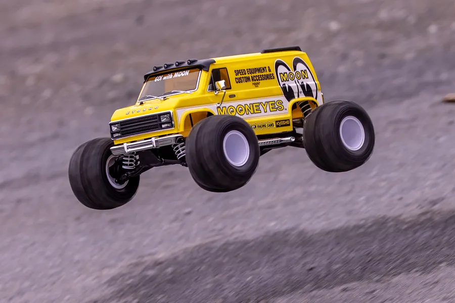 A yellow Kyosho Mad Van RC monster truck with Mooneyes branding jumping in the air over a dirt surface.