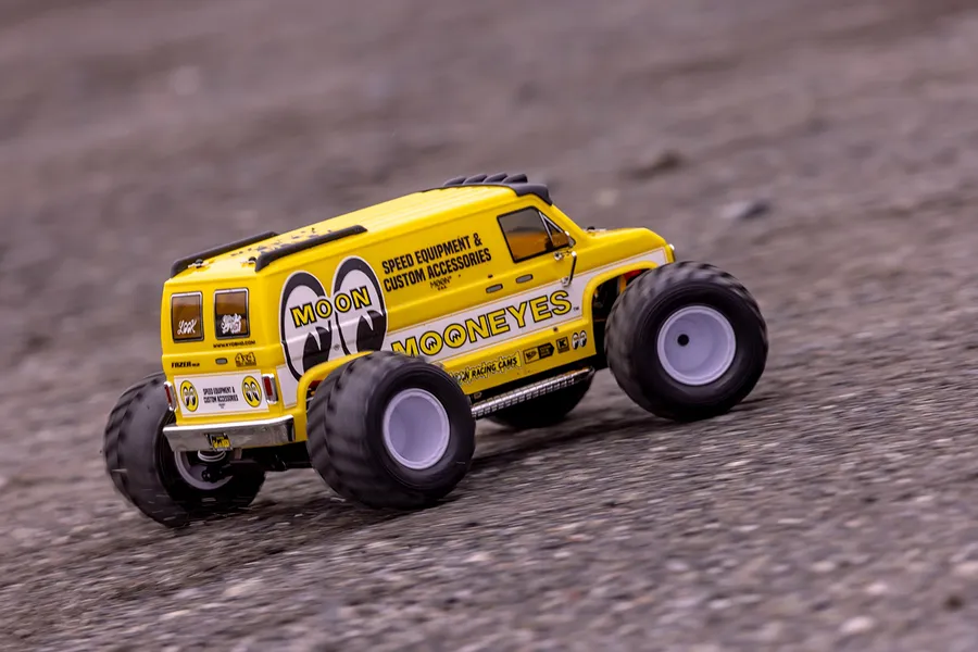 A yellow Kyosho Mad Van RC monster truck with Mooneyes branding driving on a gravel surface.