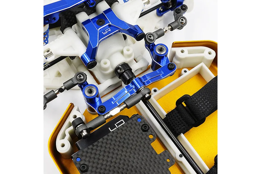 Close-up of a blue anodized aluminium steering assembly installed on an RC car chassis with white plastic and carbon fiber components.
