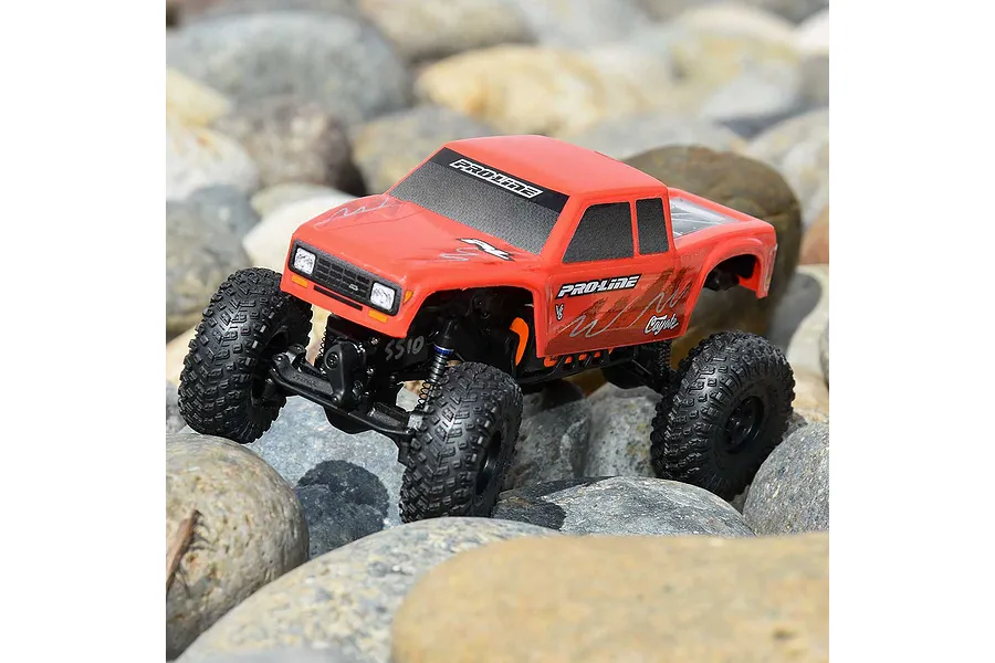 A red Pro-Line Coyote RC crawler body shell mounted on a small-scale RC chassis, driving over a bed of smooth river rocks.