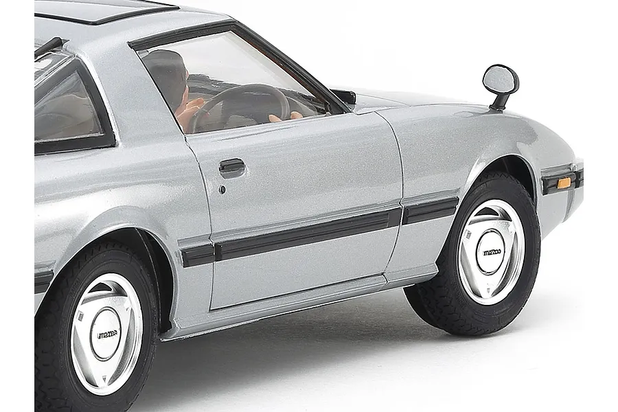Close-up side view of a silver Tamiya 1/24 scale Mazda Savanna RX-7 plastic model car showing the driver figure, wheels, and body details.
