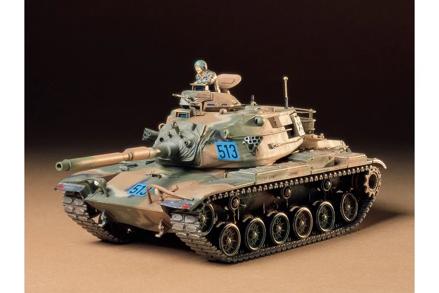A fully assembled and painted 1/35 scale Tamiya M60A3 tank model with camouflage paint and a crew figure in the turret hatch.