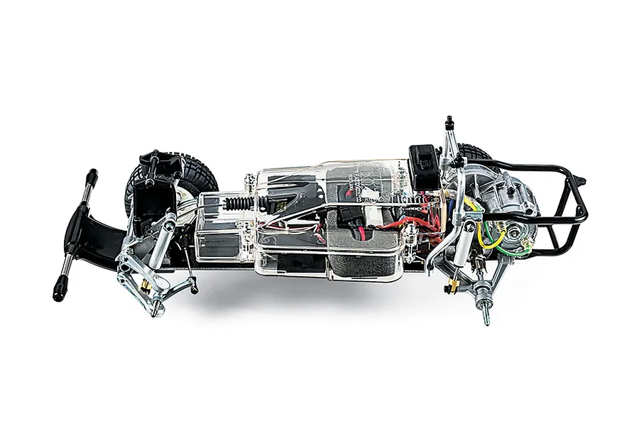A side-profile view of a Tamiya 1/10 scale RC truck chassis kit, showing the internal components, clear plastic gearbox cover, and black metal frame.