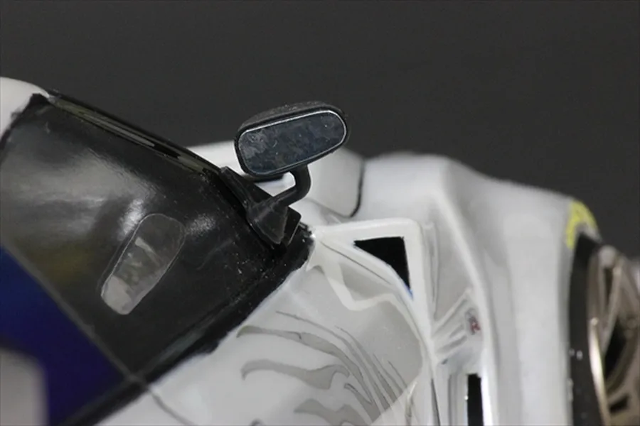A close-up view of a black racing-style side mirror mounted on a white RC car body shell.