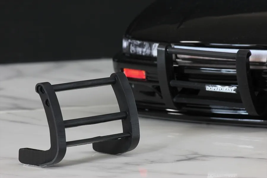 A black 1/10 scale RC grill guard sitting on a white marble surface in front of a black RC car body shell.