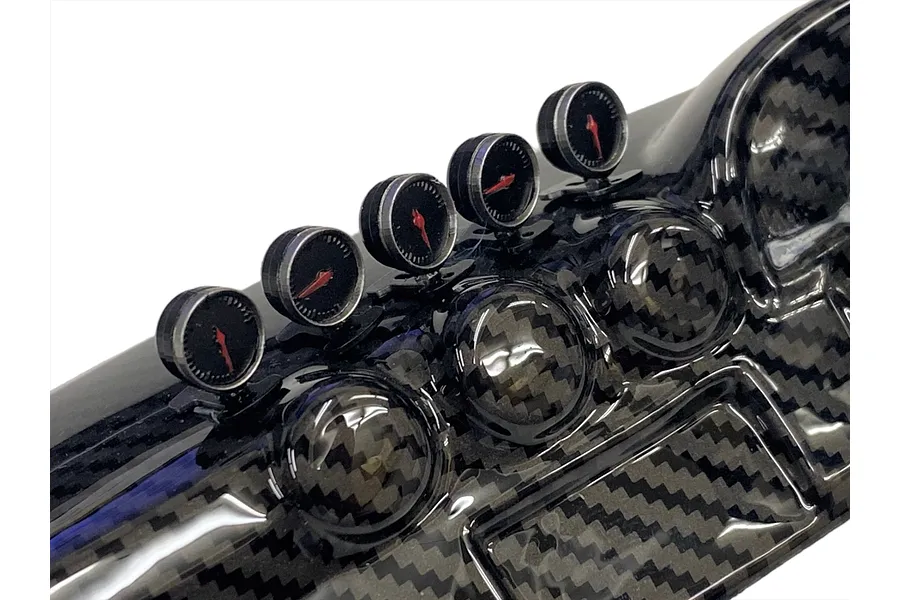 Close-up of a carbon fiber textured RC dashboard interior featuring several small round analog gauges with red needles mounted on top.