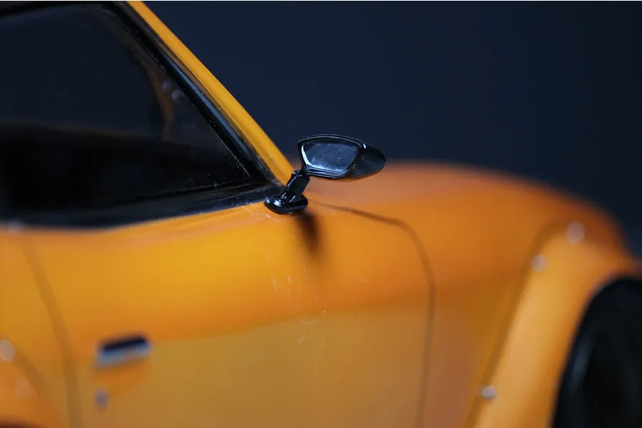 A close-up view of a black side mirror mounted on the orange body shell of a 1/10 scale RC car.