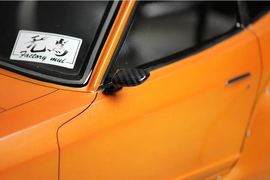 A close-up of a carbon fiber style side mirror mounted on an orange RC car body shell with a Factory mui sticker on the window.