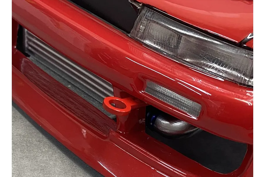 A close-up view of a red RC car front bumper featuring a small red tow hook installed below the headlight and next to the intercooler.